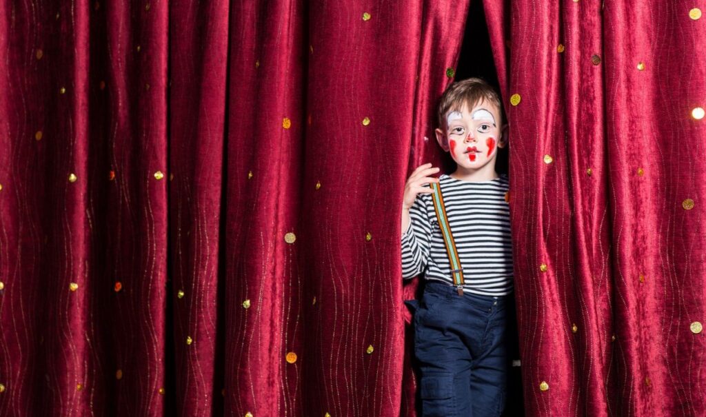 Kind als Clown sieht zwischen zwei roten Theatervorhängen hervor